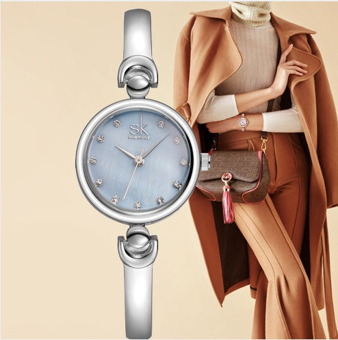 Silver watch with blue face and 'SK' brand on a beige background, with a person wearing a brown coat and holding a handbag.